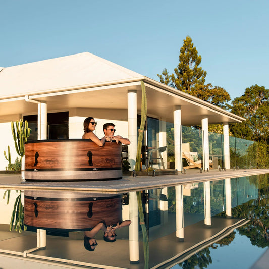Two people in a blow up hot tub by a kiwi house with a pool and trees in the background - portablespasnz