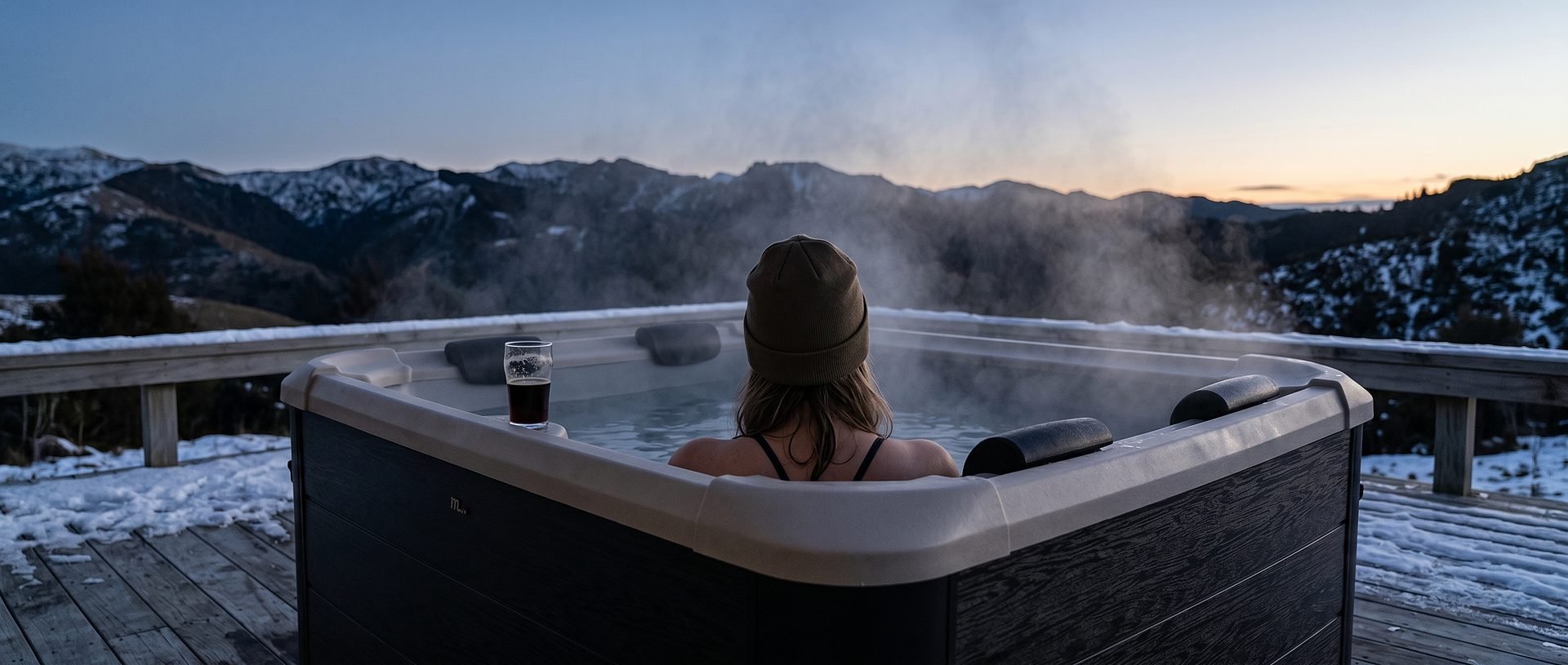 A skier sinking into a steaming portable spa pool at dusk, with a panoramic view of snow-covered Southern Alps